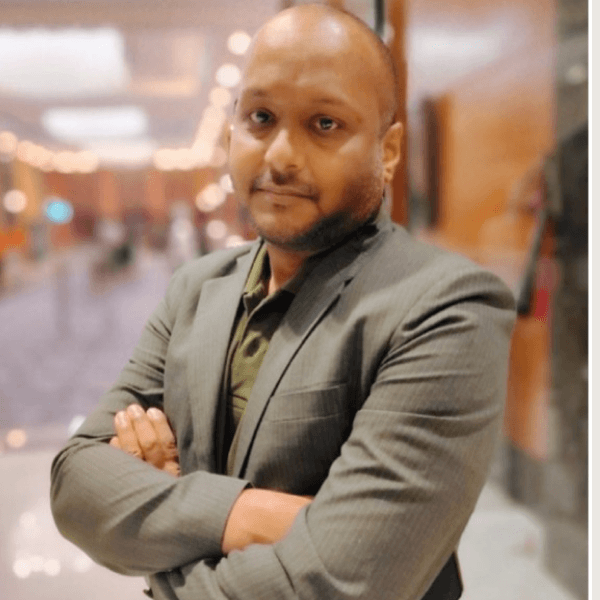 Infobip-themed graphic with a black background and decorative orange, green, and white shapes. In the center, a man stands outside a modern building looking at his smartphone while holding a coffee cup. An orange chat bubble icon appears beside him, representing messaging or RCS chat.